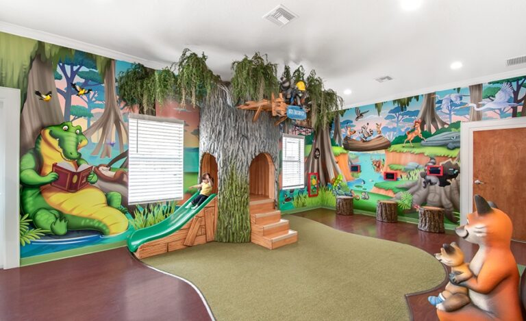 Colorful children’s playroom featuring bayou-theme mural walls of cartoon animals, a treehouse slide, and artificial grass. A crocodile reads a book amid forest-themed decorations, creating a whimsical woodland scene.