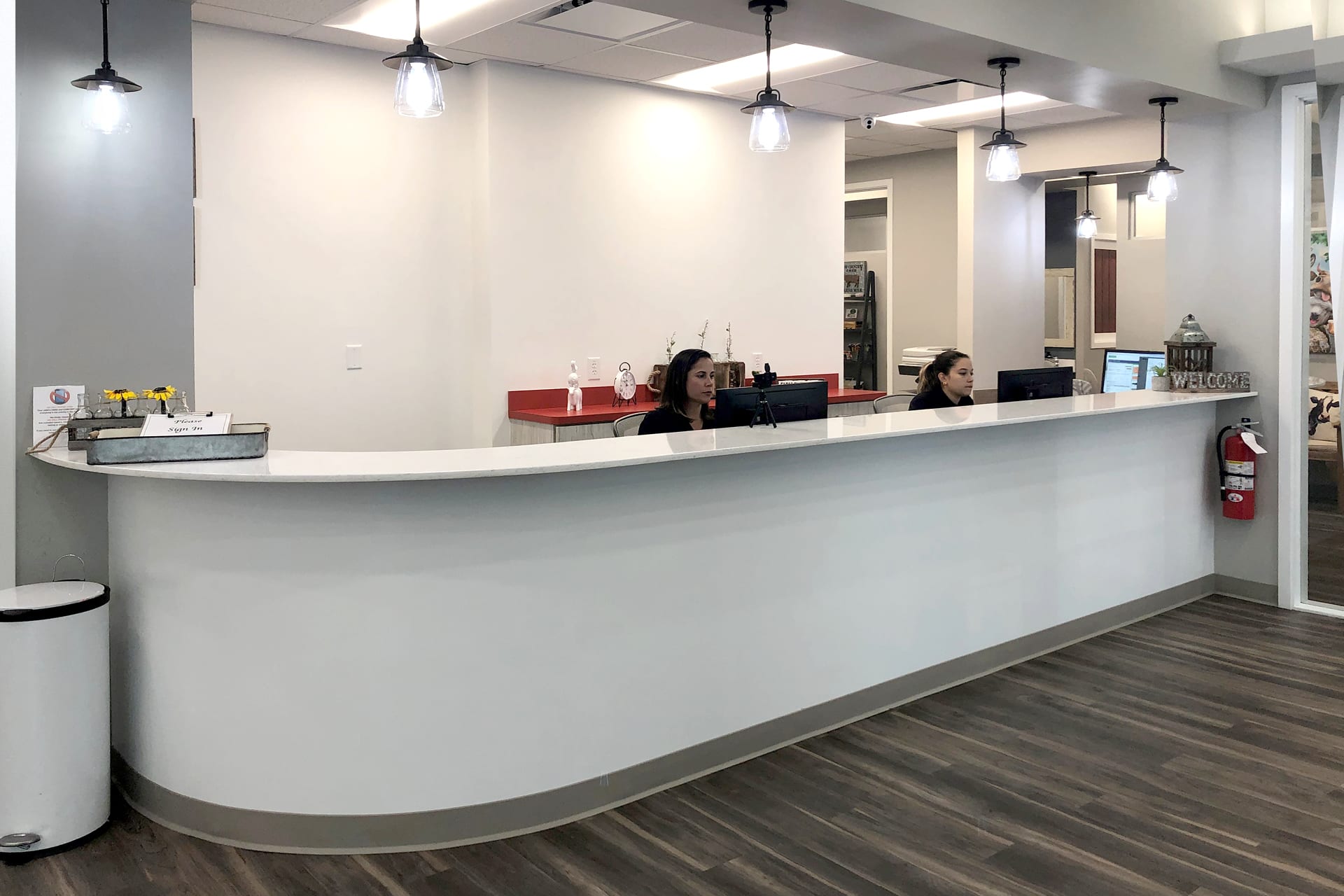 This before photo shows a modern, clean reception area with a long white counter, two receptionists at computers, pendant lights above, and wood-style flooring. A welcome sign and small decorations are on the counter.