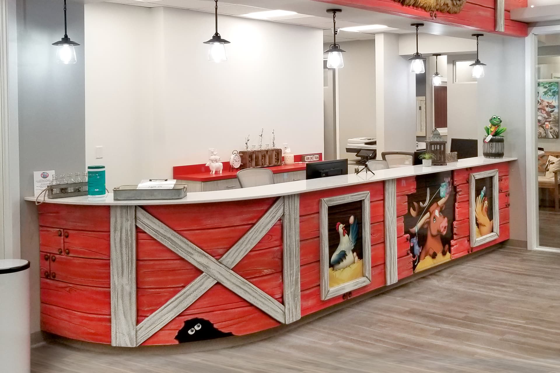 A reception desk designed to look like a red barn, with cartoon farm animals painted on the front panels, showcases a creative transformation in children's decor under hanging lights in a bright, modern office space.