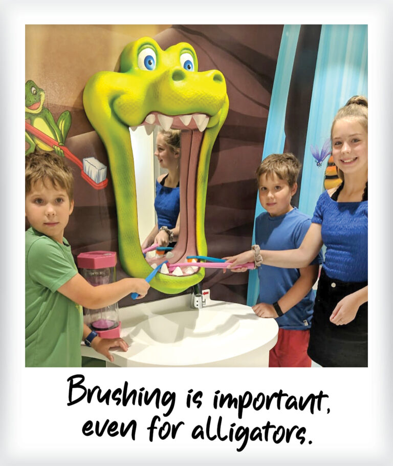 Three children stand around a sink with a large, cartoon alligator head as a mirror, surrounded by playful wall murals, pretending to brush its teeth. The text below reads, Brushing is important, even for alligators.