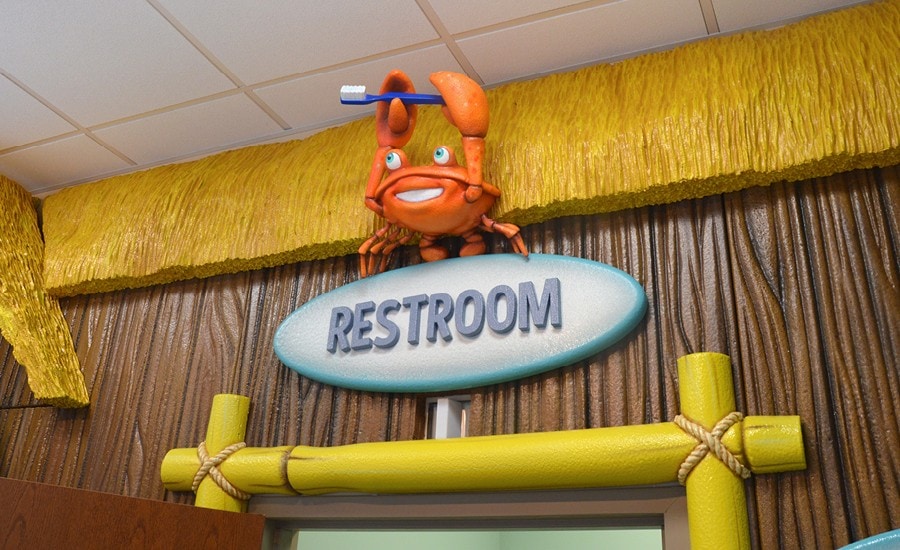 Sculpted restroom sign with a cute crab character on a thatched roof tiki hut with bamboo cladding in a pediatric dental office.
