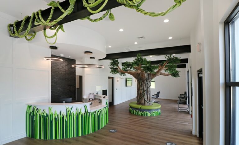A modern jungle reception area with a 3D grassy desk facade, a floor-to-ceiling tree bench centerpiece and sculpted lush green vines hanging off of a ceiling beam