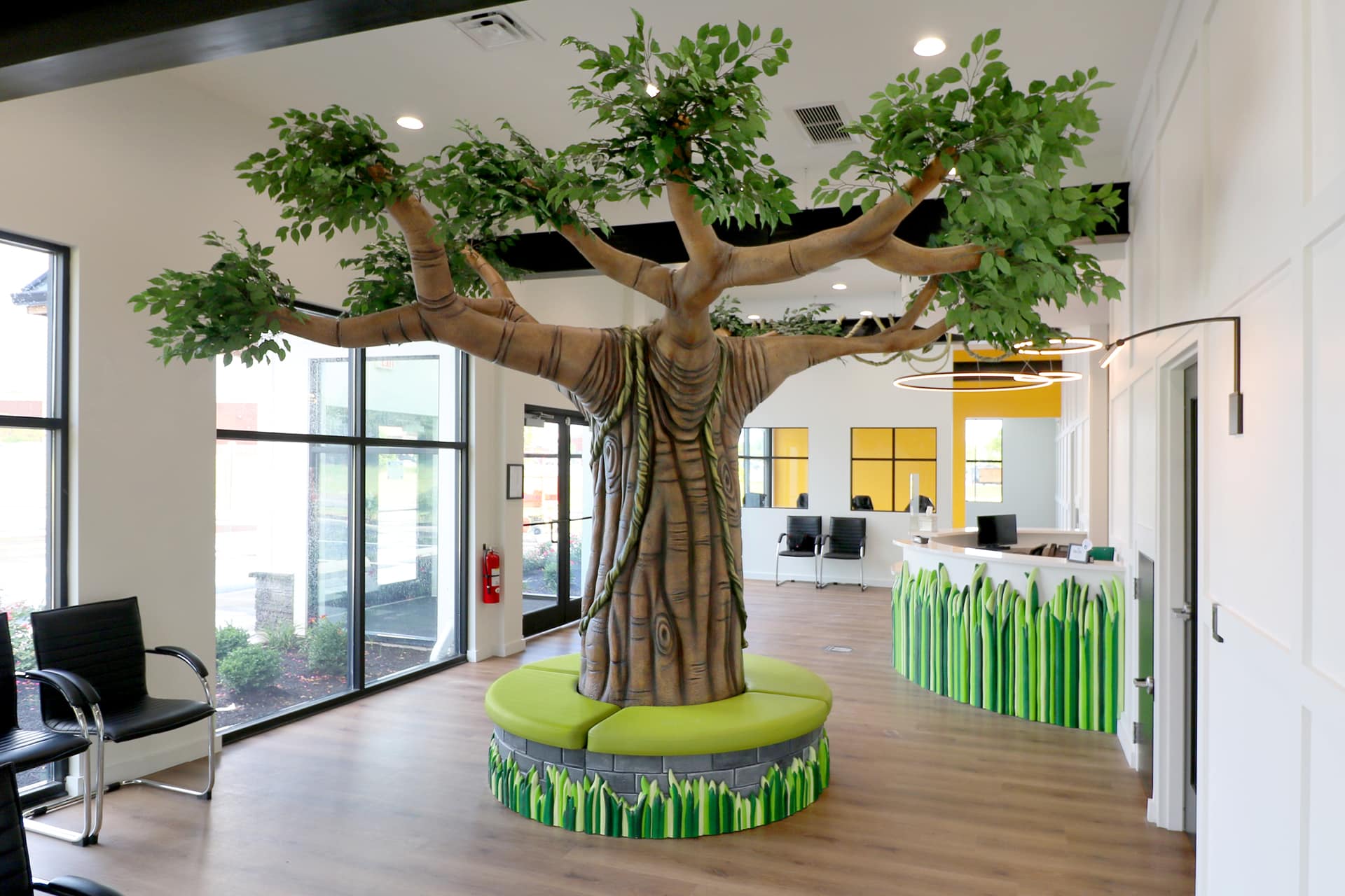 A bright, modern waiting area showcases a stunning transformation with a large artificial tree and green circular bench, perfect for children's decor, next to a reception desk adorned with playful grass shapes.