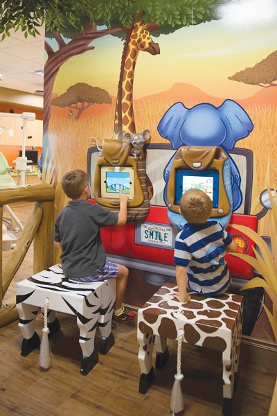 Two young boys sit on animal-shaped stools using touchscreen games set into a wall at a pediatric dental office, decorated with a colorful safari mural featuring a giraffe, elephant, and a red jeep with a SMILE license plate.