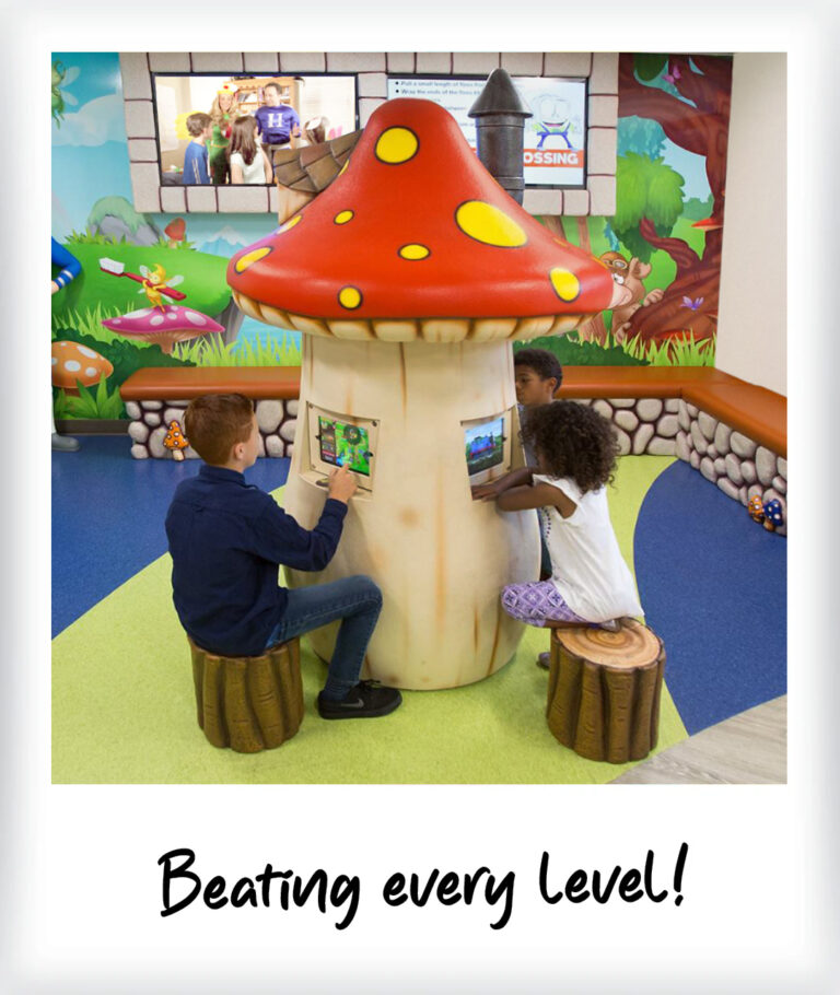 Three children sit on log-shaped stools, playing at custom video game stations embedded in a large mushroom-shaped console in a colorful, whimsical indoor play area. A caption at the bottom reads, Beating every level!.