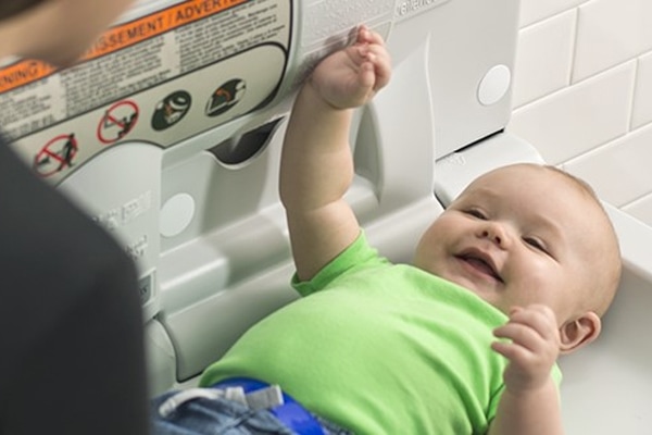 A smiling baby in a green shirt lies on a public changing table at a family church, reaching up towards the safety warning label above. White tile wall in the background.