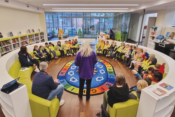 A group of young children sit in a semicircle on curved green couches in a library presentation area, listening to an adult standing in front of an alphabet-themed rug. Bookshelves and large windows are in the background.