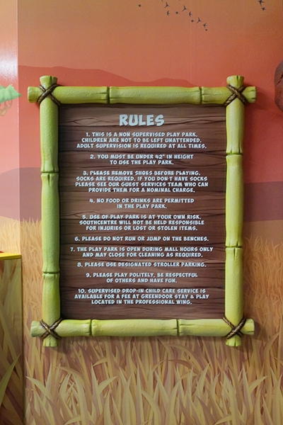 A wooden sign framed with green bamboo displays mall play area rules in white text, mounted against a colorful wall with grass illustrations. The rules outline safety guidelines and expected behavior for children and guardians.