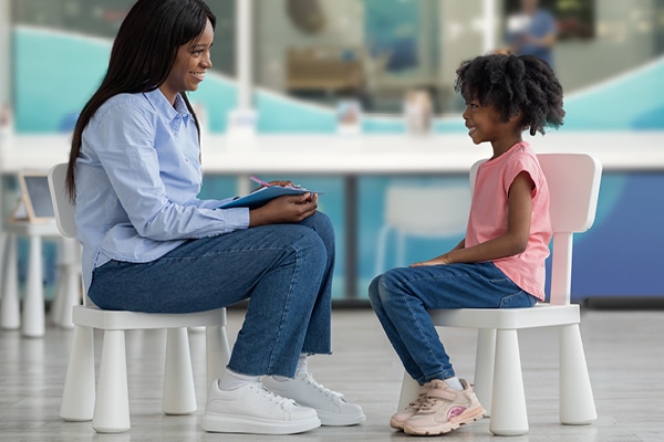 A woman and a young girl sit facing each other on small chairs indoors. The woman holds a clipboard and pen, smiling as she talks with the girl, who is also smiling. The setting appears bright and friendly.