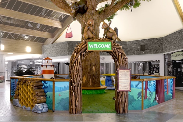 Mall play area featuring a large artificial tree, playful woodland creatures, a green Welcome sign, and colorful fences with nature-themed designs—perfect for kids to enjoy while shopping.
