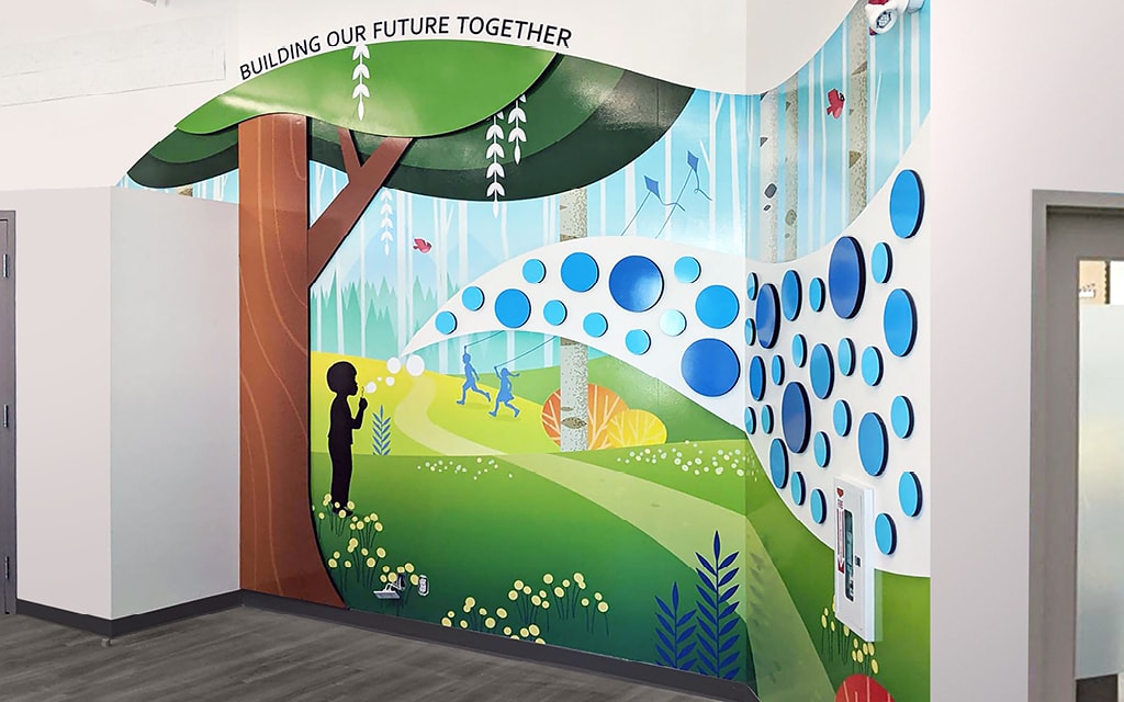 A colorful donor wall mural shows children playing in a whimsical forest with trees, plants, bubbles, and butterflies. The words Building Our Future Together are written at the top. The artwork covers two adjoining walls.