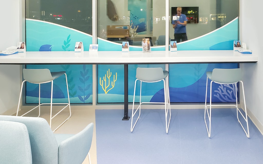 A modern waiting area with four light blue chairs at a counter facing a large window with an ocean-themed design.