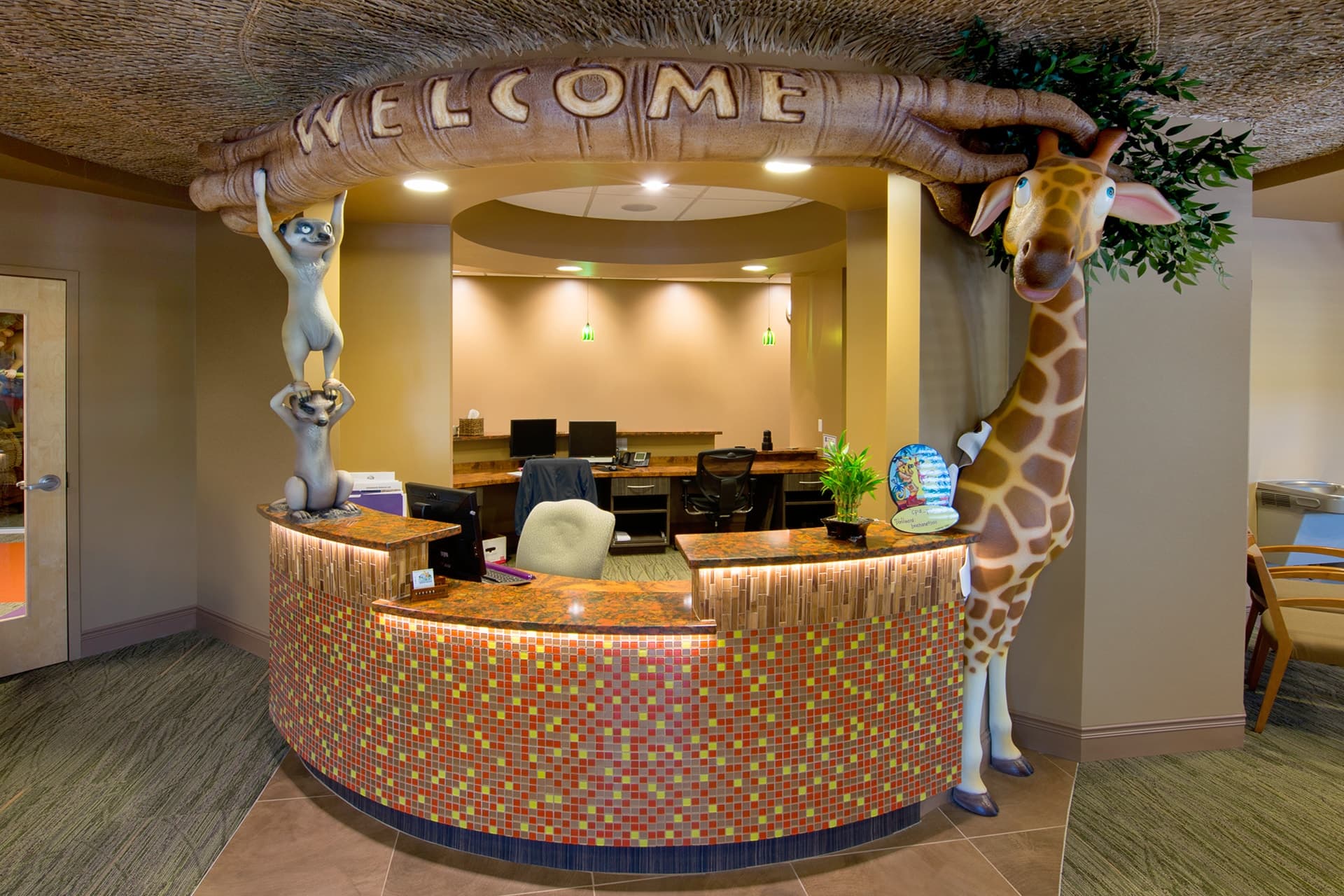 Experience a themed environment transformation: A colorful reception desk with a mosaic tile front, decorated with a large WELCOME sign held by playful animal statues, including a giraffe and lemurs, in a cheerful, jungle-themed office lobby.