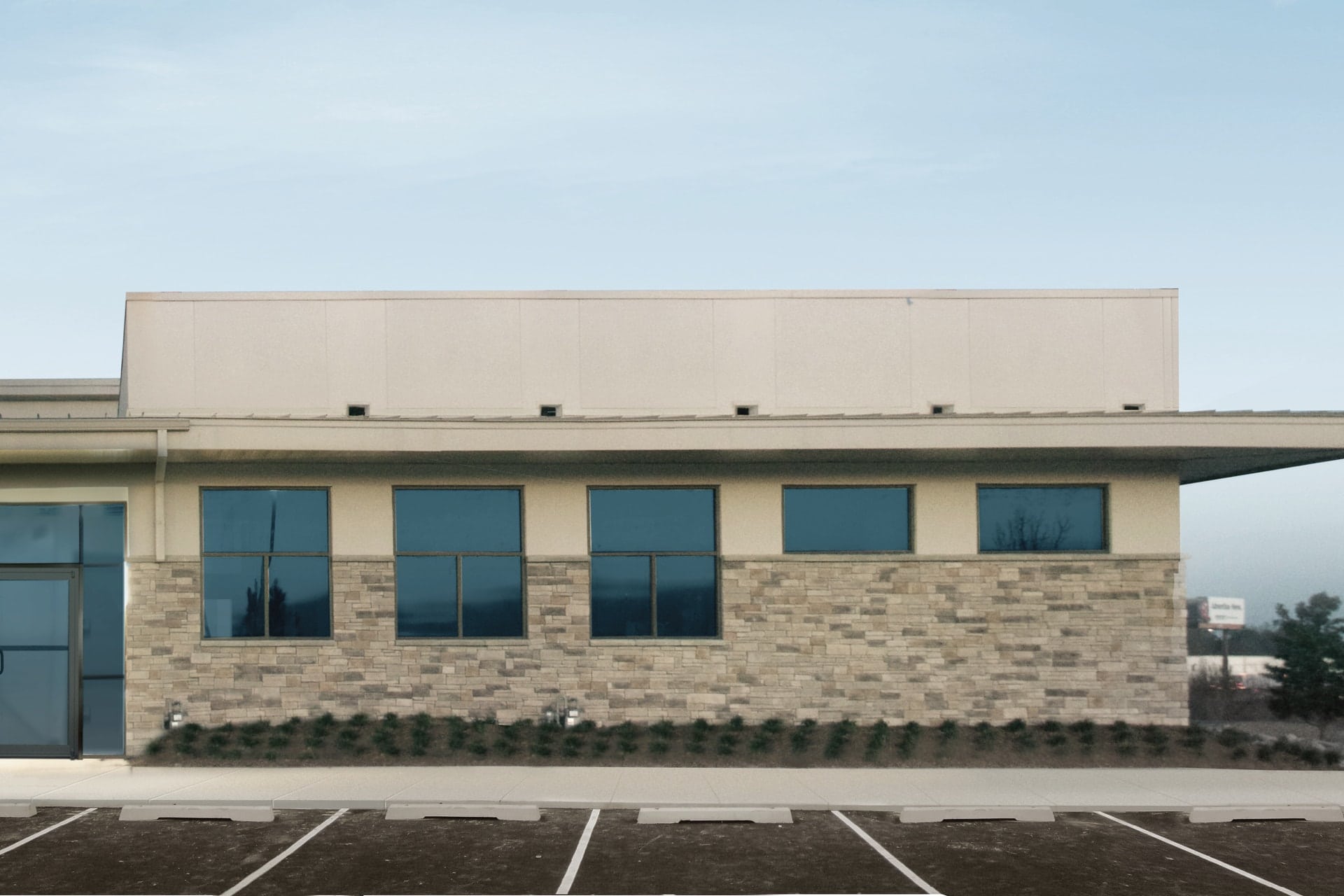 A modern commercial building with large blue-tinted windows, drab stone and stucco exterior, flat roof, and an empty parking lot in front under a clear sky—this is a boring before photo.
