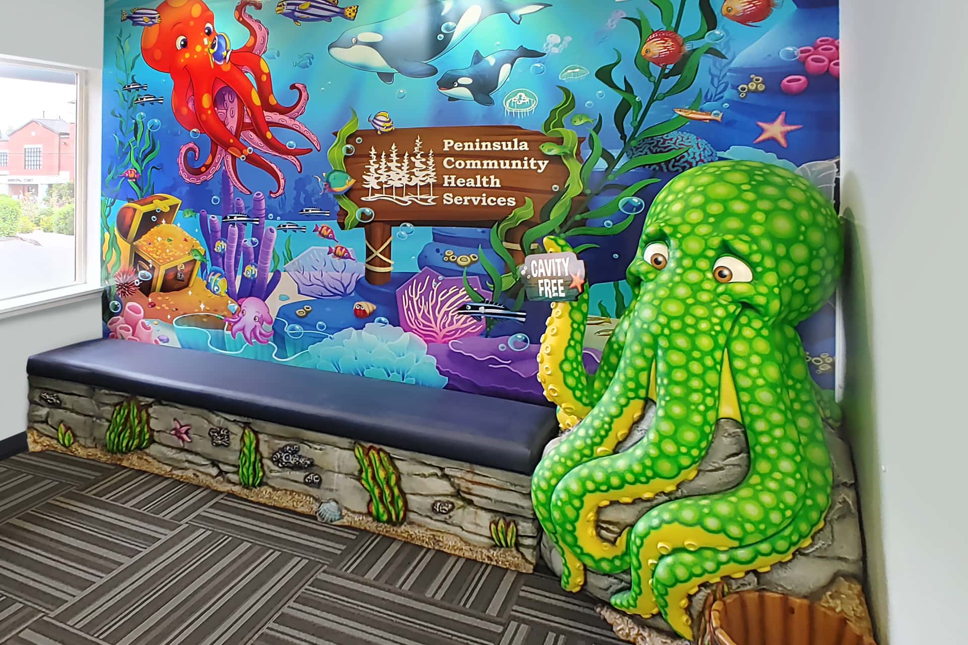 Colorful underwater-themed waiting area transformed with vibrant sea creature murals and a large green octopus sculpture holding a “Cavity Free” sign. Experience a themed environment transformation at Peninsula Community Health Services.