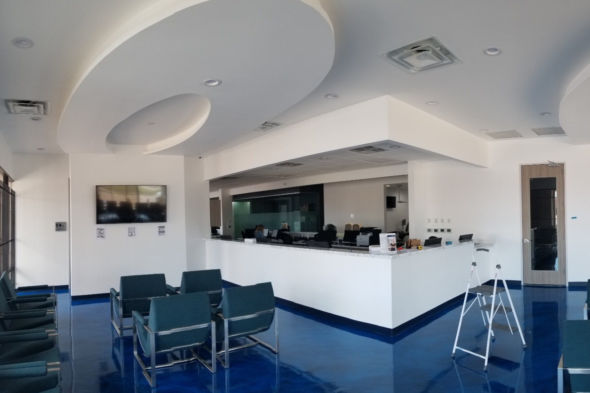 Experience a stunning themed environment transformation in this modern, spacious waiting area featuring teal chairs, a white curved ceiling, blue shiny floor, reception counter, wall-mounted TV, and a step ladder near a wooden door.