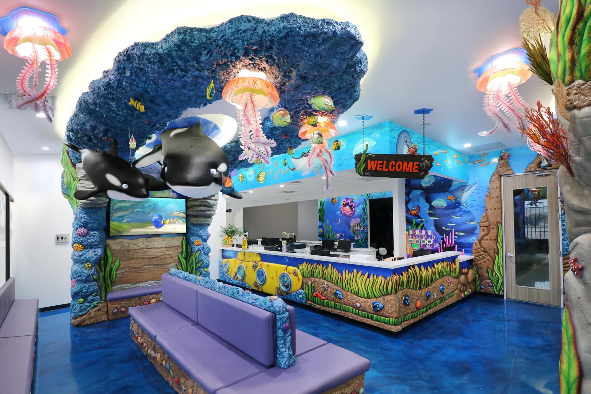Experience a remarkable themed environment transformation in this vibrant, ocean-themed reception area. Before and after, enjoy blue floors, purple seating, colorful sea life decor, and murals of whales and underwater scenes under jellyfish lights.