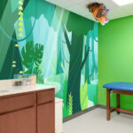Experience a themed environment transformation in this children's medical exam room, featuring a jungle-themed wall mural, green walls, a blue exam table, wooden cabinet with sink, and a tiger head decoration hanging from the ceiling.