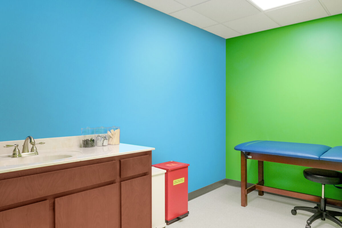A brightly lit but drab medical exam room with vibrant blue and green walls, a sink with brown cabinets, a red biohazard bin, and an exam table with blue padding and a black stool—this is a boring before photo.