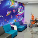 Before and after a themed environment transformation, a young boy sits next to an orange alien statue on a gray and blue couch in a modern room with a vibrant space-themed mural featuring cartoon spaceships and stars. A TV is mounted on the wall above.