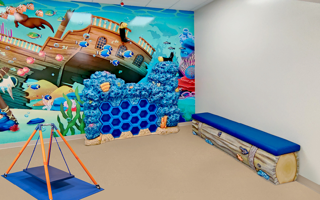 A colorful, ocean-themed pediatric therapy facility playroom with a mural of an underwater shipwreck, blue coral climbing wall, a log-shaped bench, and a small hanging swing. The room is bright, playful, and perfect for therapy activities.