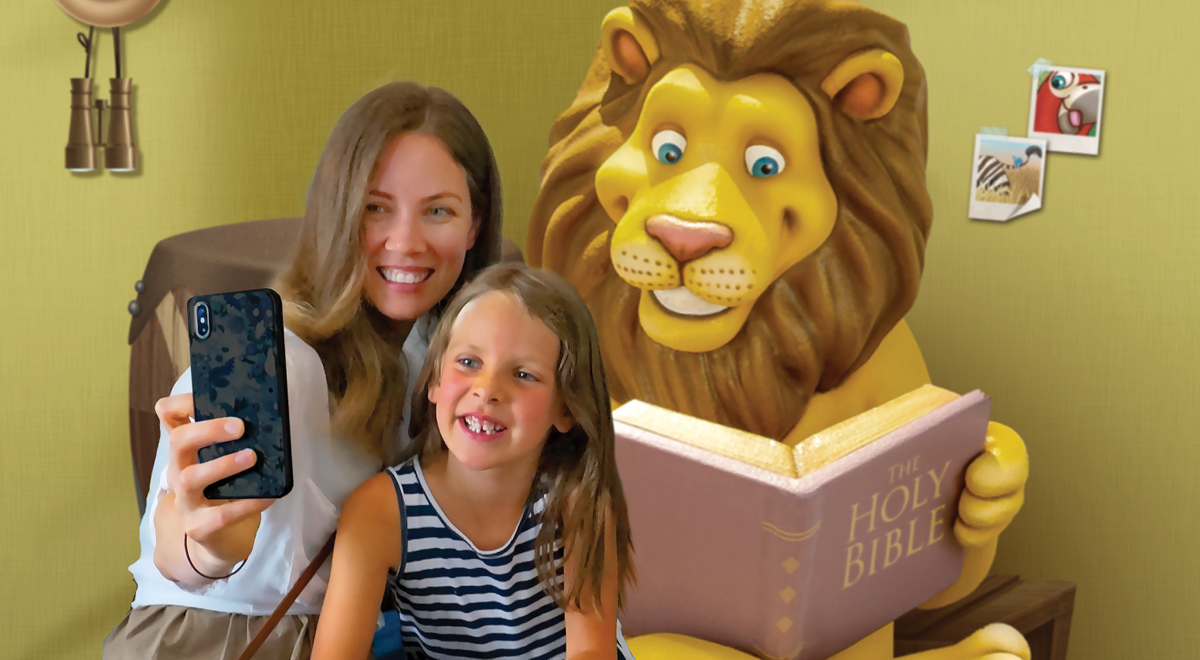 A woman and a young girl smile while taking a selfie together. Behind them, a large, friendly-looking cartoon lion holds an open Bible amid playful church decor—binoculars and animal pictures add to the cheerful scene.