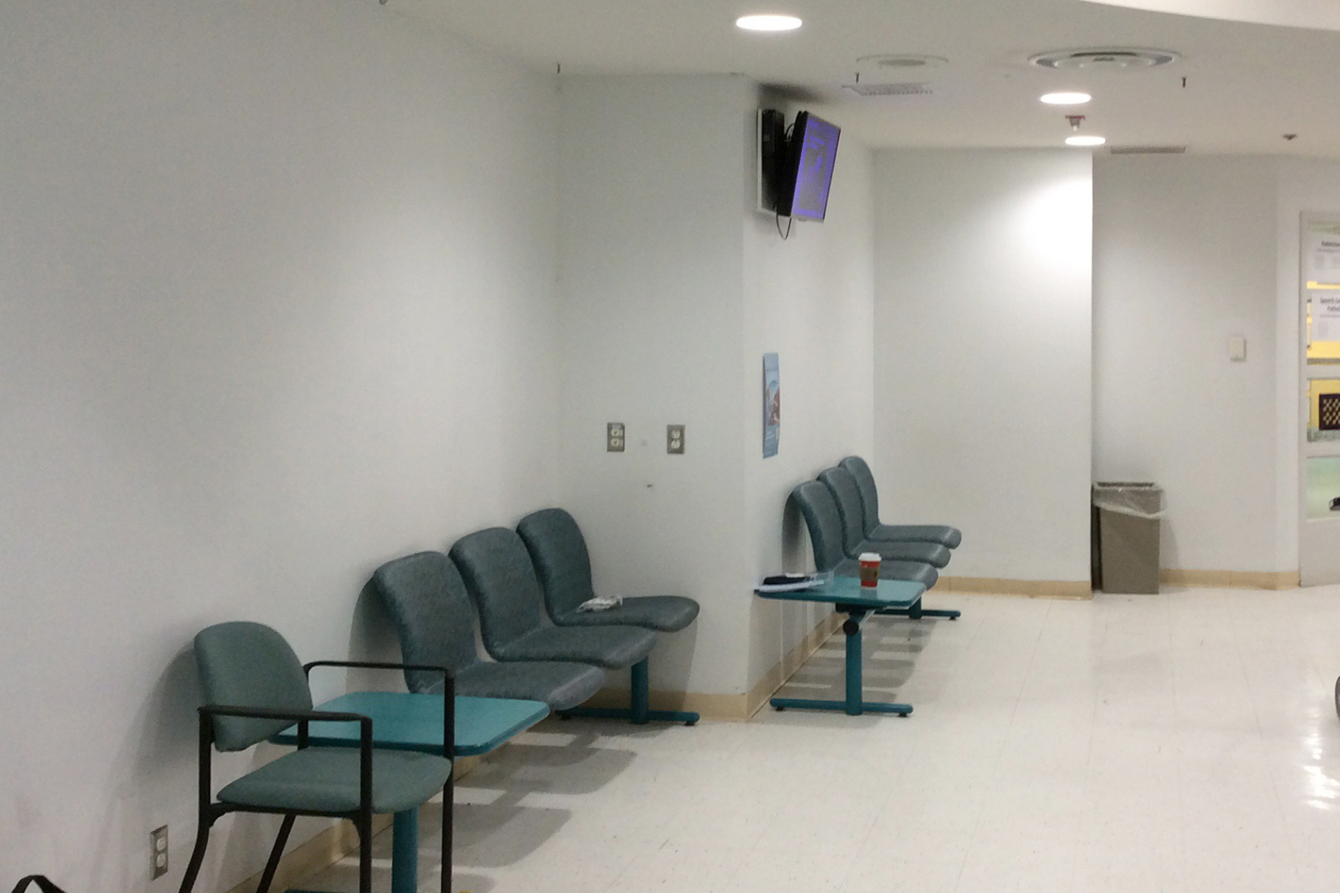 A clinical hospital waiting area with empty blue chairs lined against a white wall, a small blue table, and a trash can in the corner. The floor is shiny white, making this a boring before photo. A TV is mounted high on the wall.