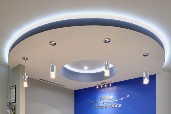 A modern orthodontic office ceiling features a large circular recessed design with soft white lighting and four hanging pendant lights. The blue and white wall below displays the Heller Orthodontics logo.