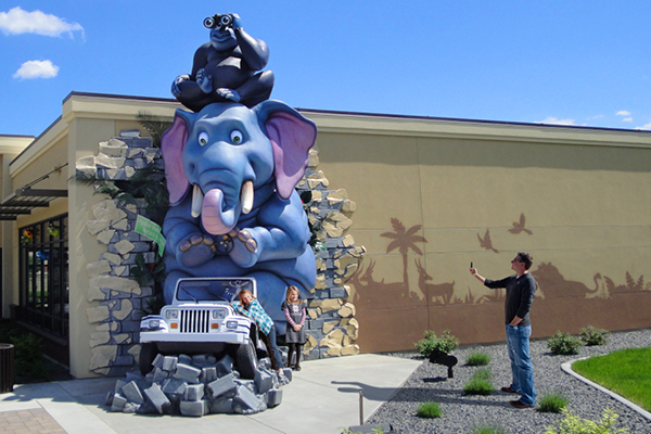 A man takes a photo of two children posing next to a large blue elephant statue sitting on a fake Jeep, with a black monkey holding binoculars on its head outside a building on a sunny day.