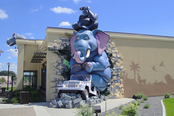 A large, cartoonish blue elephant statue appears to burst through a building wall, clutching a white jeep. A gorilla on the elephant’s head holds binoculars. The scene is whimsical, with clear skies in the background.