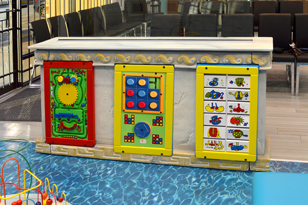 A childrens play area with interactive activity panels, featuring colorful gears, buttons, and spinning picture tiles, is set against a decorative counter. Chairs and a waiting area are visible in the background.