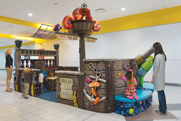 Children play on a colorful, pirate ship-themed indoor playground, decorated with sea creatures and playful designs, while two adults stand nearby watching. The scene is set in a bright, open indoor space.