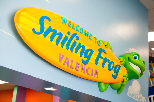 A colorful sign reads “Welcome to Smiling Frog Valencia” with a cartoon green frog holding a yellow oval sign against a blue wall with clouds.