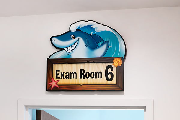 A playful wall sign for an optometrist's office with a smiling cartoon shark riding a wave above the text "Exam Room 6." The sign features a wooden design with starfish and shell details.