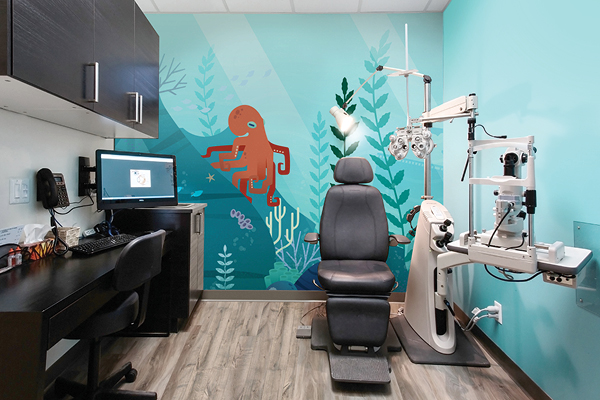 A themed optometry exam room with an underwater mural featuring an orange octopus and coral. The room includes an examination chair, optometry equipment, and a sleek desk setup.
