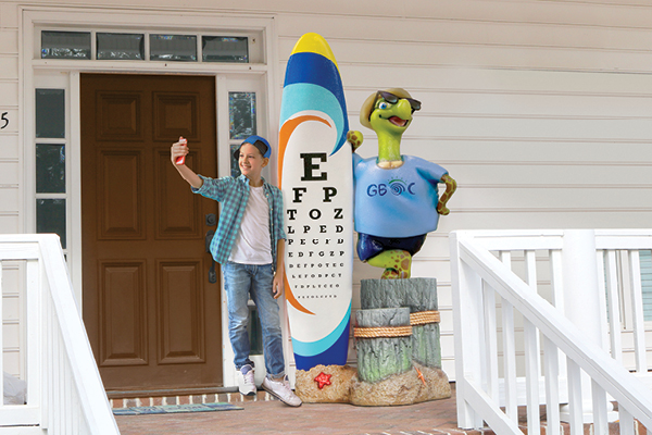  A whimsical outdoor decoration at an optometrist's office entrance, featuring a surfboard-shaped eye chart and a cheerful turtle sculpture wearing sunglasses and a t-shirt. A person takes a selfie with the decor.