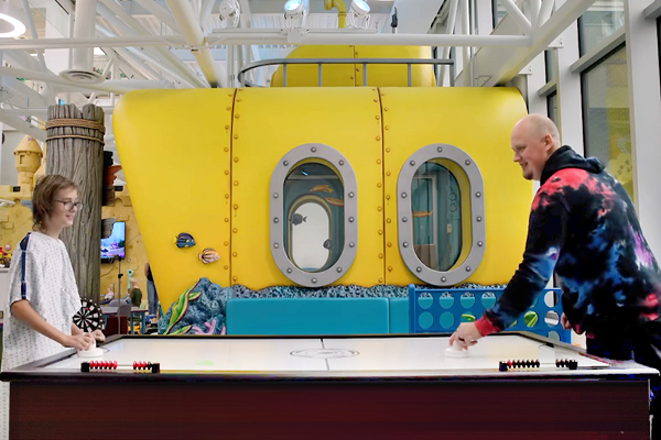 A child in a hospital gown and an adult play air hockey in front of a sculpted yellow submarine in a underwater themed family room.