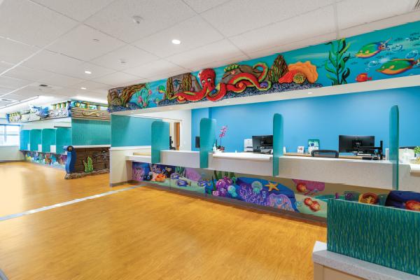 A bright, colorful Pediatric Front Desk welcomes visitors to the hospital, featuring undersea-themed wall murals with fish, an octopus, and coral, plus blue accents and a clean counter equipped with computers.