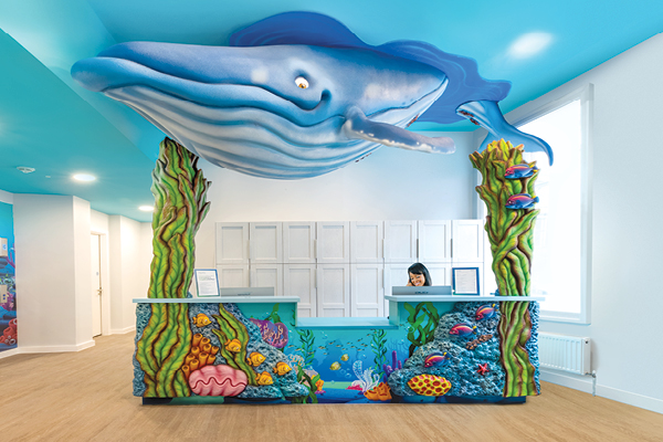 A colorful, ocean-themed reception desk designed as decor for a children's hospital features coral and sea life decorations with a large blue whale model suspended above. A receptionist sits behind the desk in a bright, modern room.