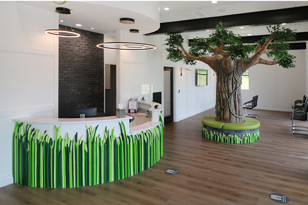 A modern pediatric front desk area features a curved desk decorated with green grass designs and a large artificial tree with a circular bench at its base. Wood flooring and black chairs along one wall create a welcoming, kid-friendly atmosphere.