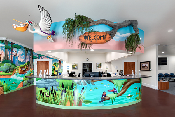 A colorful pediatric office reception area features a vibrant mural of animals and nature, a playful Pediatric Front Desk beneath a large “Welcome” sign on a faux tree branch, and a charming stork sculpture above the curved desk.
