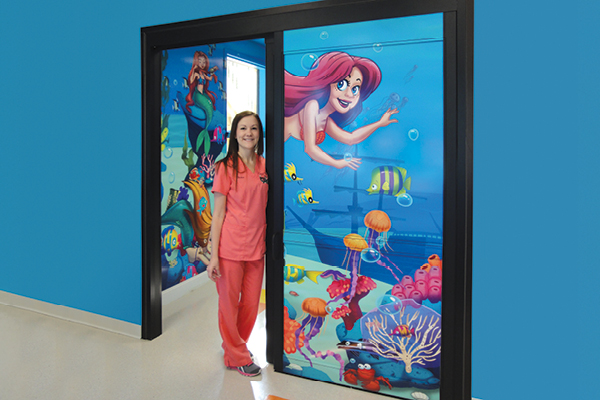 A nurse in pink scrubs stands at a sliding door adorned with hospital decor for children—colorful underwater mural featuring cartoon characters, including a mermaid—set against a bright blue wall.