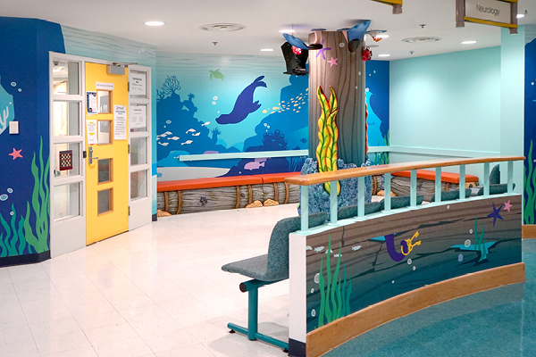 A brightly decorated hospital waiting area features an ocean theme with blue walls, murals of sea life like dolphins and fish, and colorful underwater seaweed—a cheerful decor for children's hospital visitors. Benches and railings match the aquatic design.