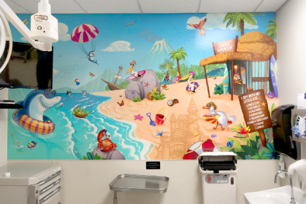 A colorful I Spy game mural of a tropical beach with animals, sandcastles, surfboards, and palm trees decorates the wall—a perfect decor for children's hospital exam rooms, creating a playful, child-friendly atmosphere.