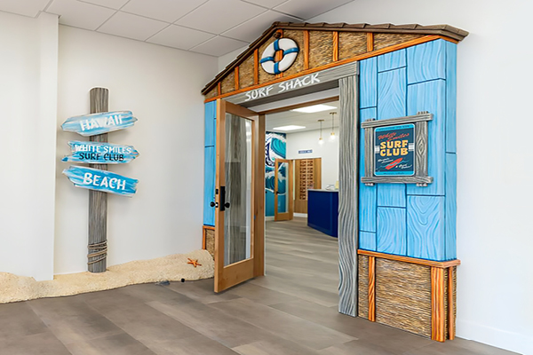 A beach-themed office entrance with a blue wooden “Surf Shack” facade, surfboard signs reading “Hawaii,” “Beach,” and “White Smiles Surf Club,” and a life preserver above the doorway. Sand and starfish decor cover the floor.