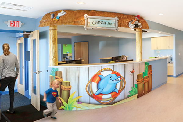 A check-in desk with a beach hut theme, featuring a thatched roof, surfboards, and a large dolphin illustration. The setting is bright and colorful.