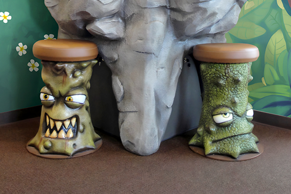 Two whimsical stools shaped like angry sugar bugs with expressive cartoon faces sit beside a large tooth carved out of rock.