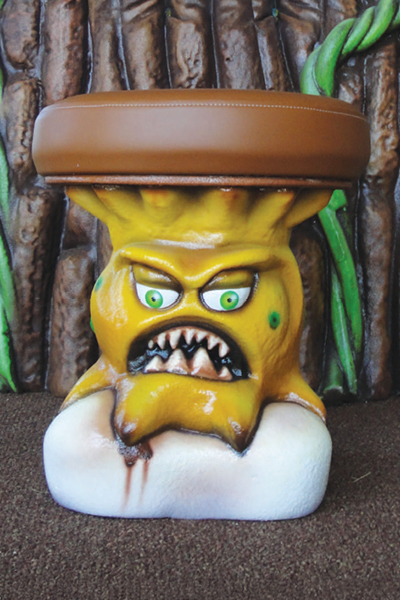 A stool designed to look like an angry yellow sugar monster with green eyes, sharp teeth, and a round brown vinyl seat sits atop a tooth in a dental office. Set against a background of tree bark and vines.