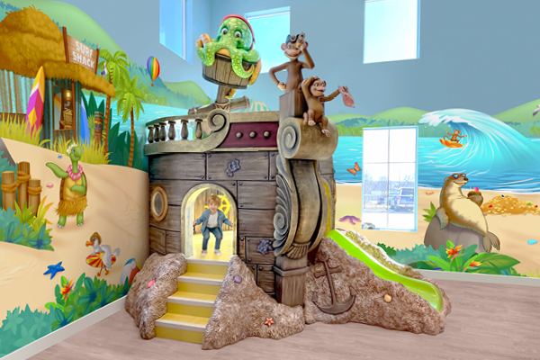 A colorful indoor play area with a pirate ship structure featuring a green pirate, monkeys, a slide, and a small entry door. The walls display a beach mural with waves, animals, and tropical decorations.