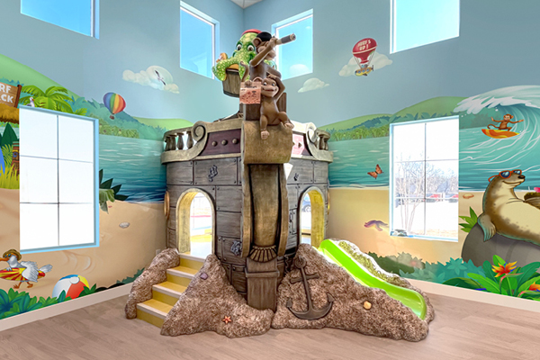 Indoor playground with a pirate ship structure featuring stairs, a green slide, and playful animal figures, set against a mural of a beach scene with sea animals, balloons, and blue sky. Pirate-themed dental office play area.